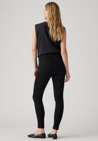 Jeans negros ajustados de cintura alta, con cinco bolsillos y un diseño elegante y ceñido. Combinados con zapatos planos negros y una blusa sin mangas negra.