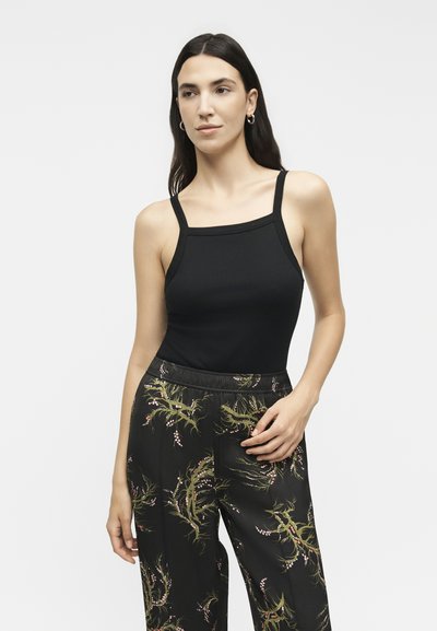 Γυναίκα με μακριά σκούρα μαλλιά που φοράει μαύρο αμάνικο τοπ και μαύρο παντελόνι με πράσινα και ροζ floral σχέδια, στέκεται μπροστά σε ένα μονόχρωμο φόντο.
