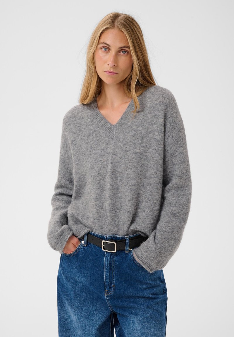 Femme blonde portant un pull gris à col en V rentré dans un jean bleu taille haute avec une ceinture noire, debout les mains dans les poches.