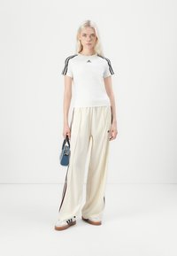 Weißes T-Shirt mit kurzen Ärmeln und schwarzen Streifen an den Schultern, kombiniert mit cremefarbenen weiten Hosen mit dunkelroten Seitstreifen. Weiße Sneakers.