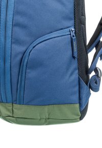 Sac à dos en tissu bleu avec un fond vert, comportant des poches zippées et une surface texturée. Bretelles réglables et détails en maille.