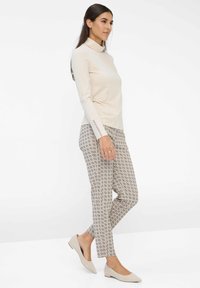 Frau in beigem Rollkragenpullover, gemusterten knöchellangen Hosen und beigen Ballerinas, die vor einem schlichten weißen Hintergrund geht.