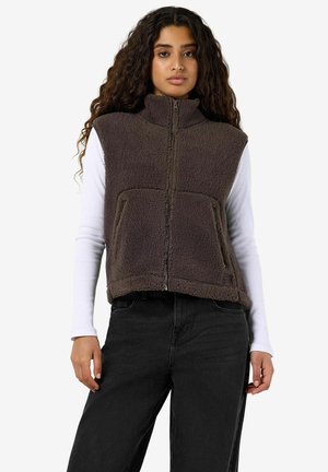 Femme aux longs cheveux bouclés portant une veste zippée en polaire marron sur une chemise blanche à manches longues et un pantalon noir, debout devant un fond blanc.