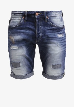 T-shirt bleu marine avec motif et poche, associé à un short en jean déchiré. Accessoires : une montre noire et des baskets bordeaux.