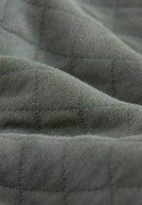 Textureret, quiltet stof i afdæmpet grøn med et gittermønster. Blødt materiale med en let glans og varierende tykkelse i sektioner.