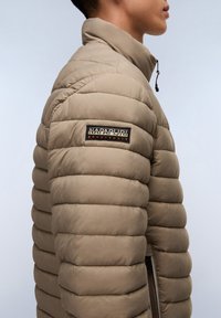 Beige pufferjakke med høj krave, kviltet design og et sort logo patch med orange bogstaver på venstre skulder.