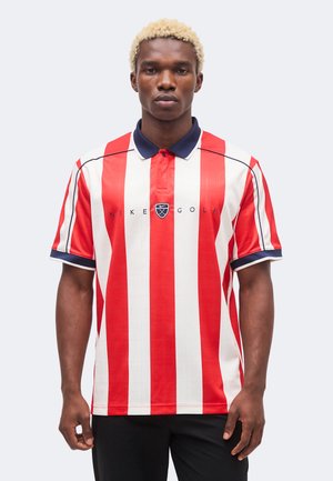 Joven con cabello rubio corto vistiendo una camiseta polo de Nike Golf con rayas verticales rojas y blancas y pantalones negros, de pie frente a un fondo claro.