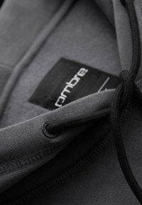 Zbliżenie na czarny materiał bluzy z kapturem z widoczną czarną sznurówką, metalowym oczkiem oraz częściowo widoczną metką wewnątrz kołnierza.