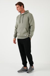 Hoodie u maslinastoj boji, s prednjim džepom i kapcima. Nositi s crnim treniranim hlačama i sivim sportskim cipelama.