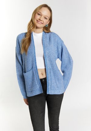 Ung kvinde smilende iført en lyseblå strikket cardigan over en hvid crop top og sorte jeans, stående mod en ensfarvet hvid baggrund.