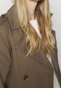 Brąco podwójnie zapięty trencz z dużym kołnierzem i błyszczącymi guzikami, charakteryzujący się gładką fakturą i zarysowaną sylwetką.