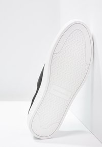 Semelle en caoutchouc blanche d'une chaussure noire avec un motif en losange et le logo "Vagabond" au centre, positionnée debout contre un coin blanc.