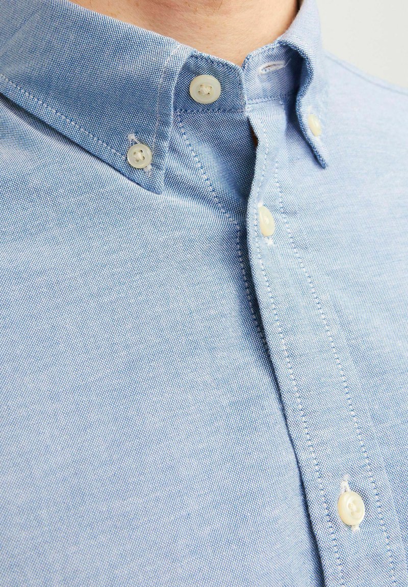 Camisa en un tono azul claro, confeccionada con una tela ligera, que presenta un cuello bien definido, botones blancos y detalles de costura finos.