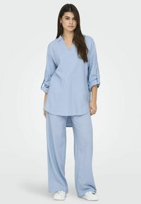 Chemise bleu clair à coupe décontractée avec col en V et manches retroussées, associée à un pantalon large assorti. Tissu lisse, détails minimaux.