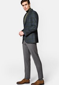 Marynarka w ciemnozieloną kratę z wełny, w połączeniu z szarymi, dopasowanymi spodniami. Całość uzupełniają brązowe skórzane buty. Pod marynarką znajduje się golf.