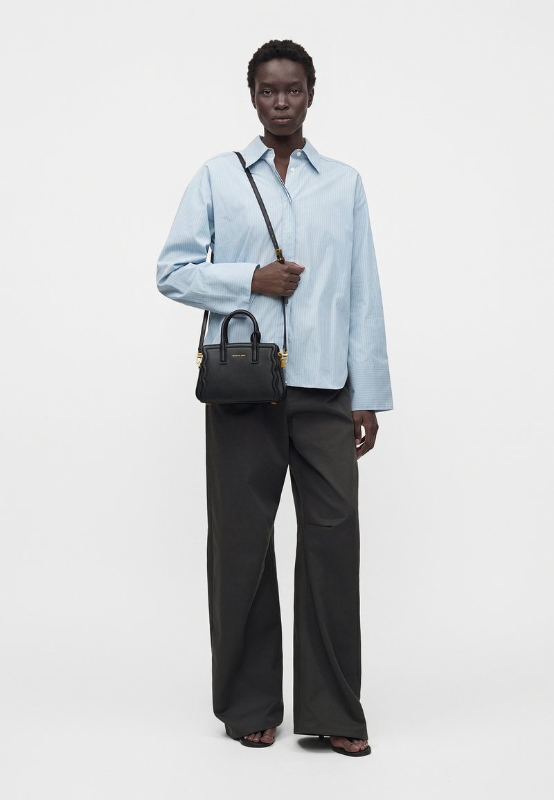Femme portant une chemise bleu clair oversize et un pantalon large noir, tenant un petit sac à main noir avec bandoulière, debout sur fond blanc.