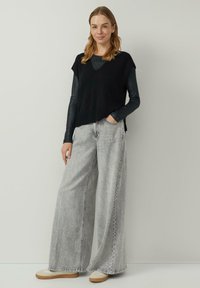 Czarna dzianinowa kamizelka z prążkowanym dołem noszona na szarym topie z długim rękawem, zestawiona z szerokimi szarymi dżinsami i beżowymi butami. Widoczne detale faktury tkaniny.