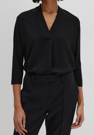 Femme portant un chemisier noir à col en V avec des manches trois-quarts, rentré dans un pantalon noir ajusté avec passants de ceinture.