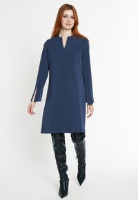 Robe bleu marine, longueur genou, avec un décolleté en V, des manches longues avec des fentes latérales. Le tissu lisse et le design simple complètent des bottes noires ajustées.