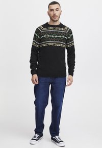 Pulover negru tricotat cu modele geometrice multicolore pe partea superioară a pieptului, purtat cu blugi albaștri și adidași negri.