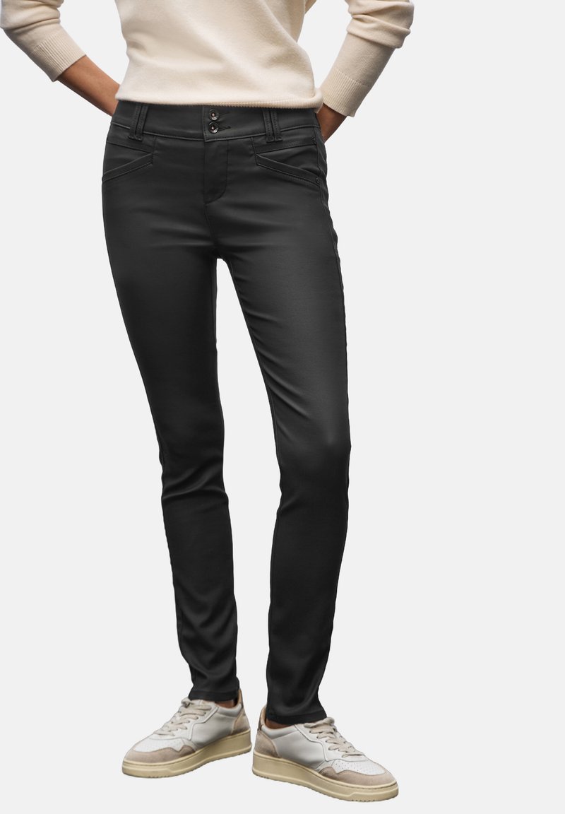 Pantalones de stretch negros con corte ajustado, que cuentan con dos bolsillos frontales, dos botones en la cintura y una textura suave. Llevados con zapatillas blancas.