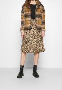 Hnědo-černý kostkovaný kabát s límcem, v kombinaci s béžovou midi sukní s leopardím vzorem a černými kotníkovými botami. Viditelné strukturované materiály.