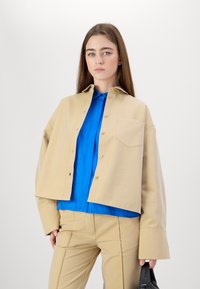 Beige cropped jacket with large sleeves, button closure, and chest pocket, layered over a bright blue shirt, paired with beige pants.