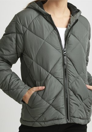 Kvinde iført en grå quiltet hættejakke med frontlynlås og hænder i sidelommerne over en hvid top.
