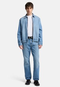 Ljusblå denimjacka med dragkedja fram, kombinerad med ljusblå jeans och en vit skjorta. Har en klassisk passform och subtila sömnadsdetaljer.