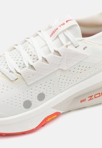 Witte sportschoen met een mesh bovenwerk, getextureerde overlays, grijze accentdoppen en een rode Nike-logo op de zool met een zichtbare dempingseenheid.