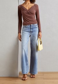Braunes langärmliges Oberteil mit Wickeldesign, hellblaue weit geschnittene Jeans und eine kleine gelbe Handtasche, kombiniert mit braunen Riemchensandalen.