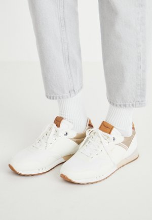 Persoon draagt witte en beige sneakers met witte sokken en lichtgrijze spijkerbroek, staande op een witte vloer.