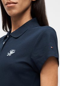 Μπλε σκούρο πόλο με υφασμάτινη υφή, με λευκό κεντημένο λογότυπο και μικρό τρίχρωμο σήμα στο μανίκι.