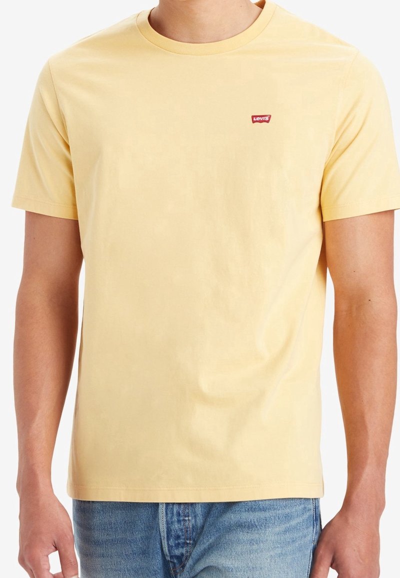 T-shirt en coton jaune à manches courtes, décolleté rond et petit logo rouge sur la poitrine gauche. Porté avec un jean en denim bleu.