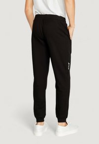 Black sweatpants with a tapered design, elastic waistband, side pockets, and white branding detail along the left seam. Paired with white shoes.