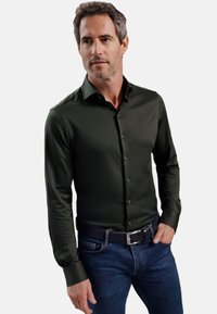 Donkergroene shirt met lange mouwen, een gladde textuur en knoopsluiting, gecombineerd met blauwe jeans en een zwarte gevlochten riem.