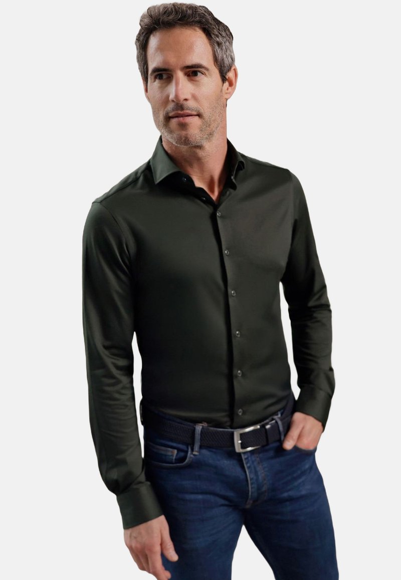 Donkergroene shirt met lange mouwen, een gladde textuur en knoopsluiting, gecombineerd met blauwe jeans en een zwarte gevlochten riem.
