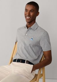 Polo gris à manches courtes avec un col ; présente un logo brodé bleu sur la poitrine. Porté avec un pantalon de couleur claire et une ceinture tissée.