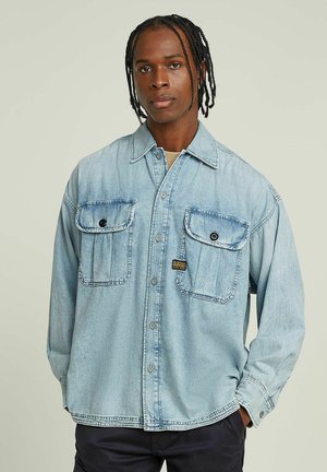 Mann mit geflochtenem Haar, der ein hellblaues Jeanshemd mit Knöpfen und zwei Brusttaschen sowie schwarze Hose vor einfachem Hintergrund trägt.