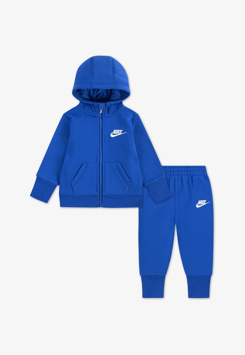 Chándal azul de Nike que incluye una sudadera con capucha y cremallera con bolsillos y pantalones a juego de corte ajustado, ambos con el logo de Nike en blanco. El material es suave y elástico.