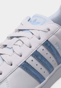 Λευκό αθλητικό παπούτσι Adidas Superstar με ανοιχτό μπλε σουέτ ρίγες, κορδόνια και λογότυπο στη γλώσσα, που δείχνει πλευρικές και πάνω λεπτομέρειες.