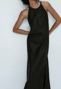 Schwarzes, ärmelloses Kleid aus einem glatten, leichten Stoff. Es hat einen hohen Ausschnitt und einen seitlichen Schlitz, der einen fließenden Fall präsentiert.