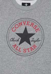 Sudadera gris con un logo redondo que presenta una estrella negra, texto rojo "CONVERSE", "Chuck Taylor" y "ALL STAR" con un diseño de borde punteado.