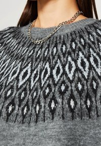 Gros plan sur un pull en tricot gris avec un motif géométrique en losanges noir et blanc et un collier en chaîne argenté porté par une personne.