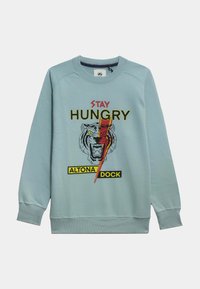 Sudadera azul claro con un gráfico de tigre, texto "STAY HUNGRY" y etiqueta "ALTONA DOCK" en amarillo. Textura suave con puños acanalados.