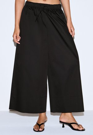 Žena v čiernych širokých nohaviciach s elastickým pásom na šnúrku a čiernych sandáloch na podpätku, nohavice sú orezané v oblasti pása.