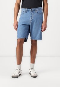 Lichtblauwe denim shorts met een ontspannen pasvorm, vijf zakken en een ruwe zoom. Gecombineerd met witte sneakers met zwarte accenten en grijze zolen.
