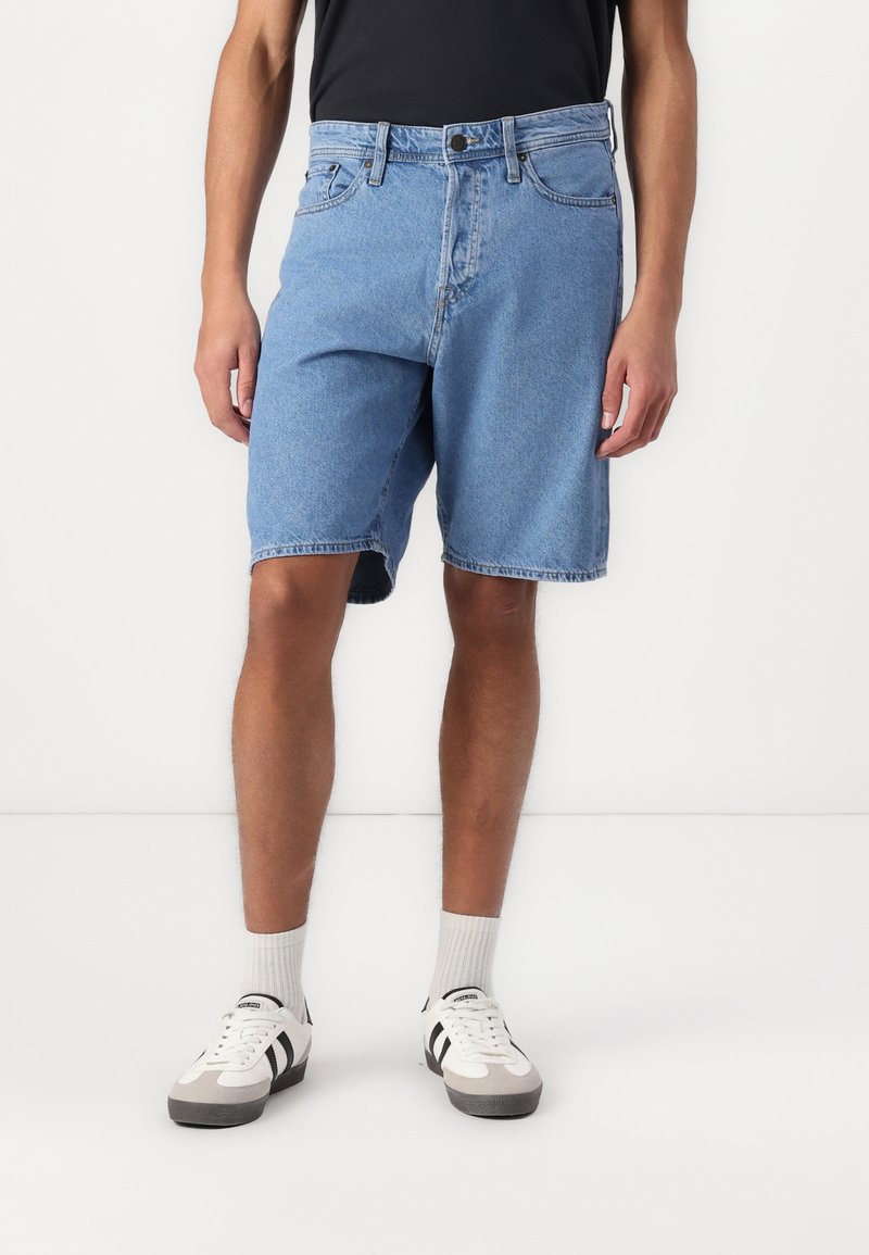 Lichtblauwe denim shorts met een ontspannen pasvorm, vijf zakken en een ruwe zoom. Gecombineerd met witte sneakers met zwarte accenten en grijze zolen.