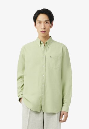 Joven con una camisa de botones verde claro con un bolsillo en el pecho y pantalones gris claro, de pie frente a un fondo blanco liso.