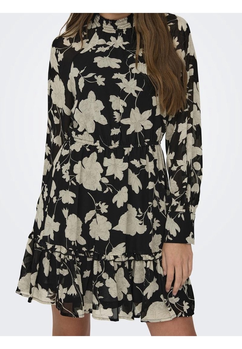 Robe noire à manches longues avec imprimé floral beige, col montant, taille froncée et ourlet à volants portée par une personne aux longs cheveux bruns.